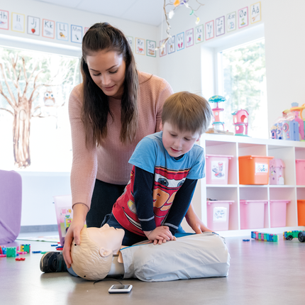 En kvinna demonstrerar HLR-docka Little Junior QCPR medan ett barn lär sig hjärt-lungräddning i en färgglad miljö.